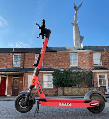 Photo of a Voi-branded red e-scooter parked in front of a house with a shark sculpture in the roof